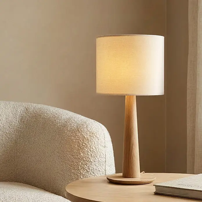 Home Sweet Home Lampenschirm Bling - Ø x H: 20 x 17 cm, Pure White, Baumwolle, Rund - Eine moderne Tischlampe mit hellem Holzfuß und zylindrischem Stoffschirm, spendet warmes Licht auf einem Holztisch neben einem cremefarbenen Sessel.
