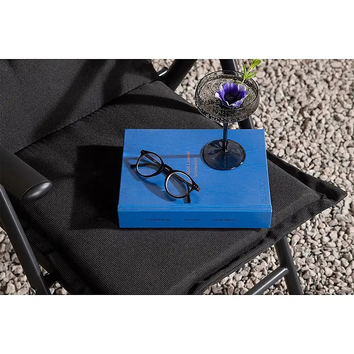 Ebuy24 Sitzauflage Cus - 1 Stk., L x B x H: 46 x 116 x 6 cm, Fasern, Schaum, Polyester., Schwarz - Schwarzer Klappstuhl aus Stoff mit einem blauen Buch, Brille und Cocktailglas darauf, auf Kies platziert.