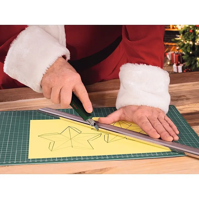 Bosch Adventskalender - Gesamtumfang: 44 -tlg. - Eine Person im Weihnachtsmannkostüm schneidet mit einem grünen Bosch Cuttermesser und Lineal einen Stern aus gelbem Papier auf einer Schneidematte.
