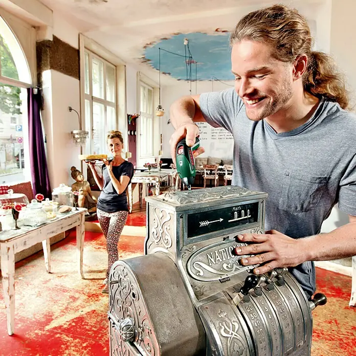 Bosch Atornillador de batería IXO Basic - 3,6 V, 1 batería, 1,5 Ah, 4,5 Nm, 10 pzs. - Un hombre repara una caja registradora antigua en un café con un taladro inalámbrico, mientras una mujer sirve pasteles.