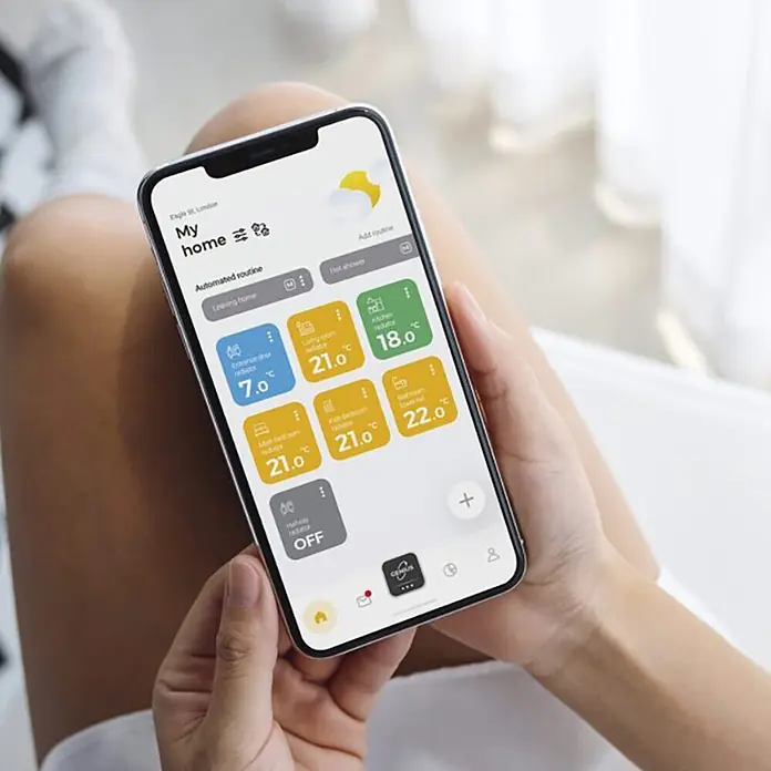 Termostato digital Genius - An x Al: 8,6 x 8,6 cm, Negro - Una persona sostiene un smartphone que muestra una aplicación de hogar inteligente para controlar la temperatura y las rutinas.