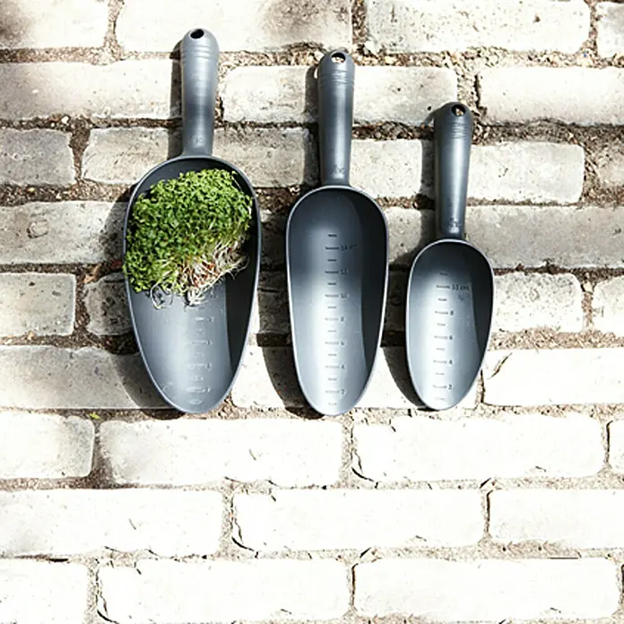 Elho Green Basics Pala - Ancho: 26 cm, Negro - Tres cucharadas medidoras grises para jardinería cuelgan de una pared de ladrillo, una llena de plantas.
