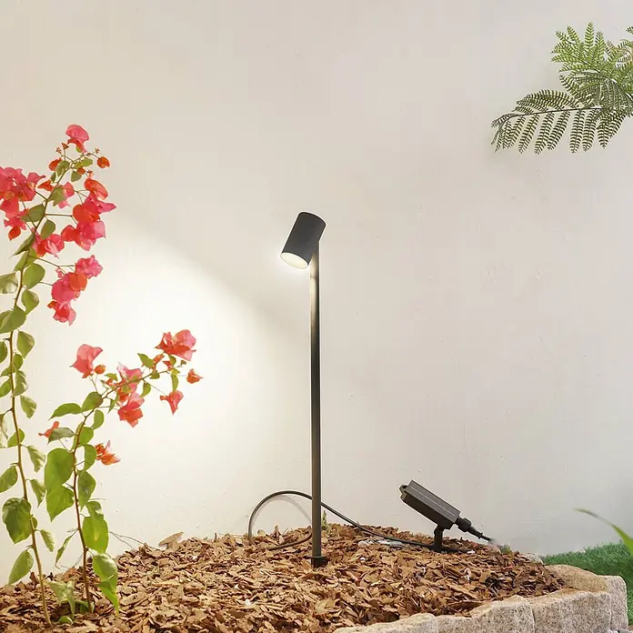 Lindby LED Solar Gartenlampe Erdspieß in schwarz matt, Erdspießlampe -  - Dunkler Außenstrahler mit Solarpanel, der eine Gartenmauer und Pflanzen in einem Mulchbeet beleuchtet.