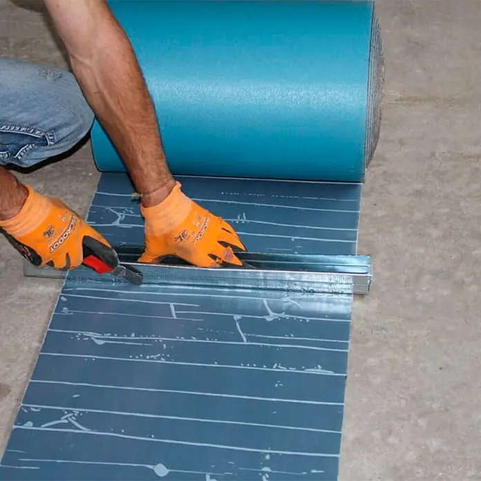 Bur2000 Cinta de insonorización Air Bur Band 500 - 12 m x 50 cm x 5 mm - Persona cortando una membrana impermeabilizante azul oscuro con un cúter y una regla metálica de un rollo.