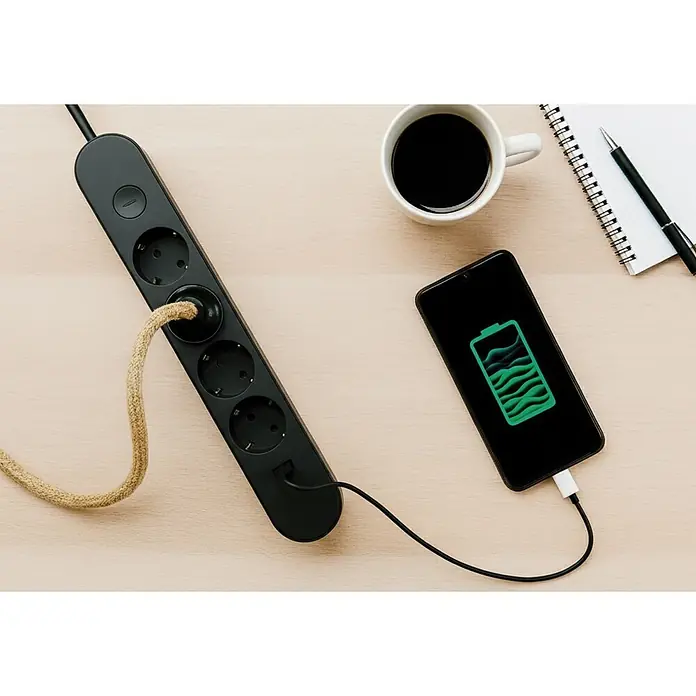 Base de enchufe múltiple Small Device Essential 4 - Negro, Longitud del cable: 1,5 m, Con interruptor - Regleta negra con cable trenzado de tela, cargando un smartphone. Una taza de café y una libreta están también en el escritorio.