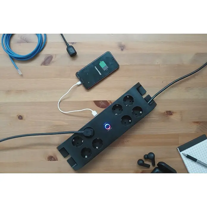 Base de enchufe múltiple Office 8 - Negro, Longitud del cable: 2 m, Con interruptor, Número de enchufes Schuko: 8 ud. - Regleta negra con ocho enchufes Schuko y un puerto USB, que carga un teléfono. Sobre un escritorio de madera con otros accesorios tecnológicos.