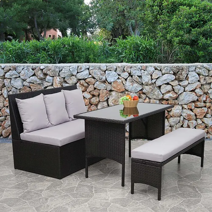 Mendler Loungemöbel-Set HWC-G16 - 3 -tlg., Polyrattan, Schwarz, Kissen hellgrau, Schwarz - Dunkelbraune Rattan-Gartenmöbelgarnitur mit hellgrauen Kissen, bestehend aus Sofa, Glastisch und Bank, auf einer Steinterrasse.