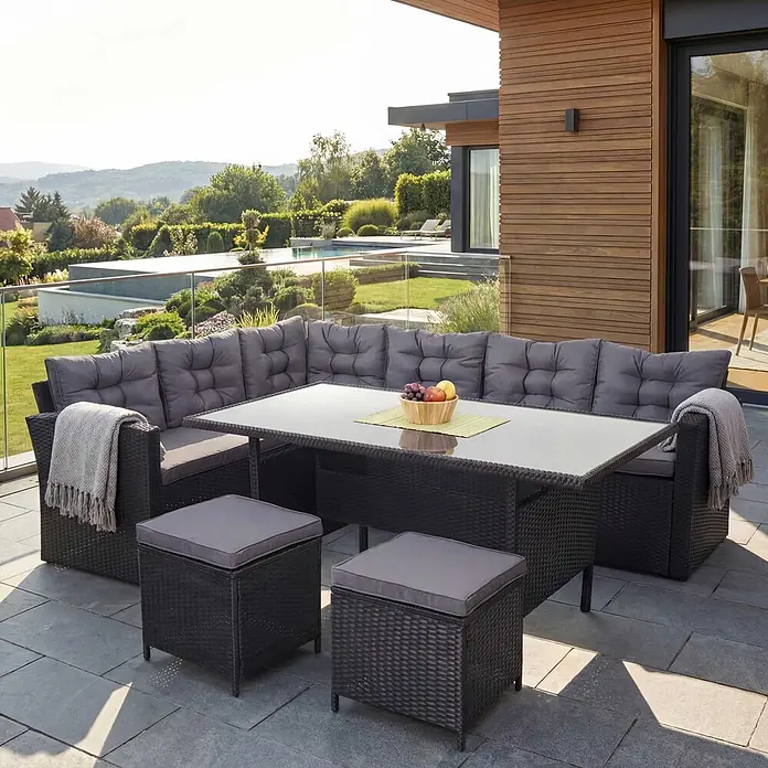 Mendler Loungemöbel-Set HWC-A29 - 4 -tlg., Polyrattan, Kissen dunkelgrau, mit 2x Hocker, Schwarz, dunkelgrau - Dunkelgraues Rattan-Gartenmöbelset mit L-Sofa, Glastisch und zwei Hockern auf einer modernen Terrasse.