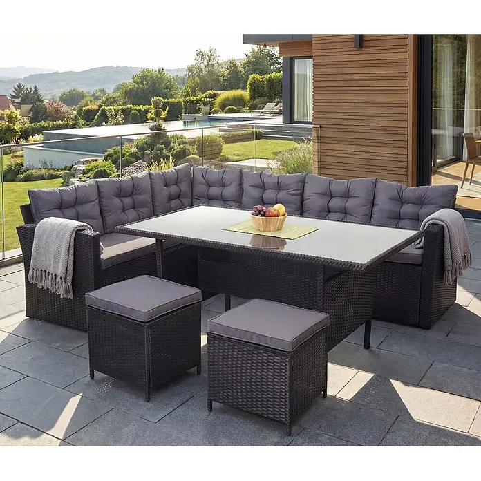 Mendler Loungemöbel-Set HWC-A29 - 4 -tlg., Polyrattan, Kissen dunkelgrau, mit 2x Hocker, Schwarz, dunkelgrau - Dunkelgraues Rattan-Ecksofa-Set mit Glastisch und zwei Hockern, grauen Polstern, auf einer Terrasse.