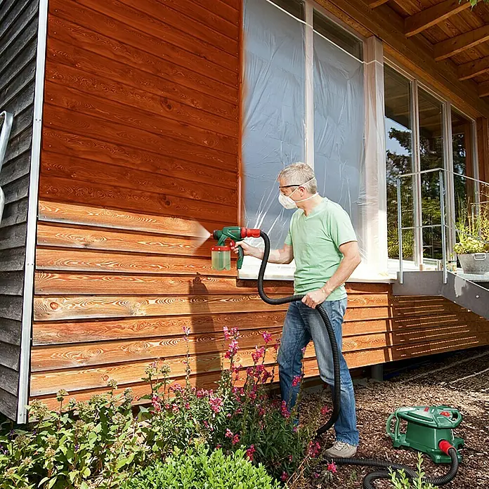 Bosch Farbsprühsystem PFS 5000 E - Geeignet für: Alle Farbarten, Nennaufnahme: 1.200 W, 1.000 ml - Mann lackiert Holzfassade mit Farbspritzpistole. Das Gerät ist grün und rot, während der Mann eine Maske und Jeans trägt.