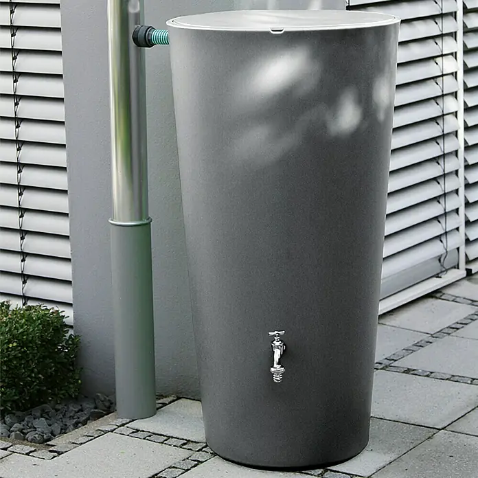Regenspeicher Rainbowl Flower - 150 l, Anthrazit - Regentonne in Anthrazit mit einem Wasserhahn zur Gartenbewässerung, neben einem Fallrohr auf Steinplatten platziert.