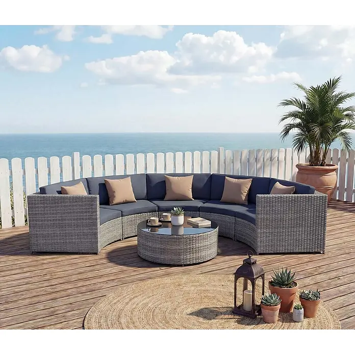 Mendler Loungemöbel-Set HWC-E17 - 6 -tlg., Polyrattan, Grau, Polster dunkelgrau, Kissen beige, Grau, dunkelgrau - Graues Rattan Outdoor-Sofa in Kurvenform mit blauen Polstern, beigen Kissen und rundem Glastisch auf einer Terrasse mit Meerblick.
