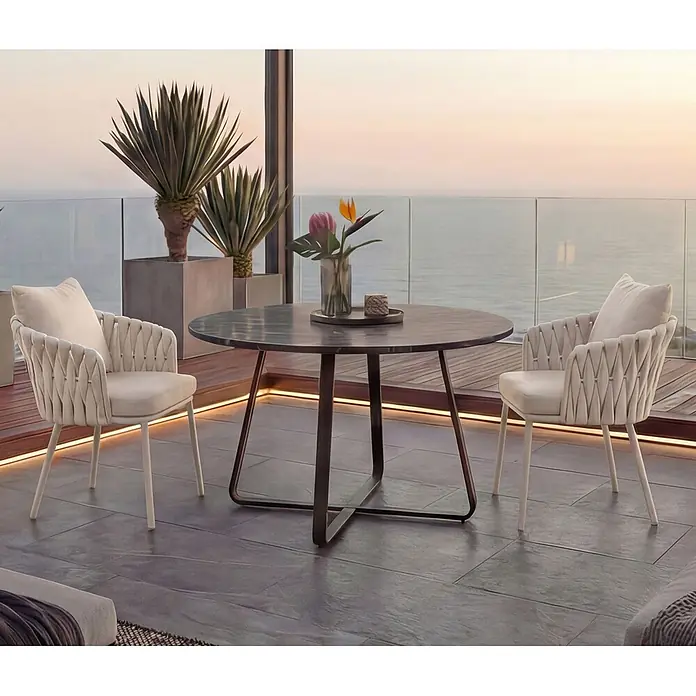 Mendler Gartenstuhl-Set HWC-P89 - 2 Stk., Metall, Creme, B x T x H: 56 x 57 x 71 cm - Outdoor-Essgruppe mit dunklem runden Tisch und zwei hellbeigen geflochtenen Sesseln auf einem Balkon mit Meerblick.