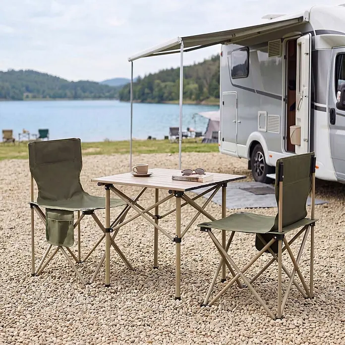 Mendler Campingset HWC-Q39 Tisch, 2x Stuhl, Metall Stoff - grün -  - Khakifarbene Klappstühle und ein hellbrauner Tisch auf einem Kiesplatz, vor einem Wohnmobil an einem See.