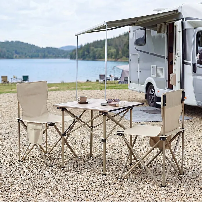 Mendler Campingset HWC-Q39 Tisch, 2x Stuhl, Metall Stoff - creme -  - Beiger Klapptisch und zwei passende Klappstühle im Freien neben einem Wohnmobil an einem See.