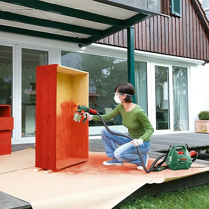 Bosch Farbsprühsystem PFS 3000-2 - Geeignet für: Alle Farbarten, Nennaufnahme: 650 W, 1.000 ml - Eine Frau lackiert mit einer Bosch PFS 3000-2 Farbspritzpistole ein Holzregal im Freien, um ihm einen neuen, frischen Look zu verleihen.