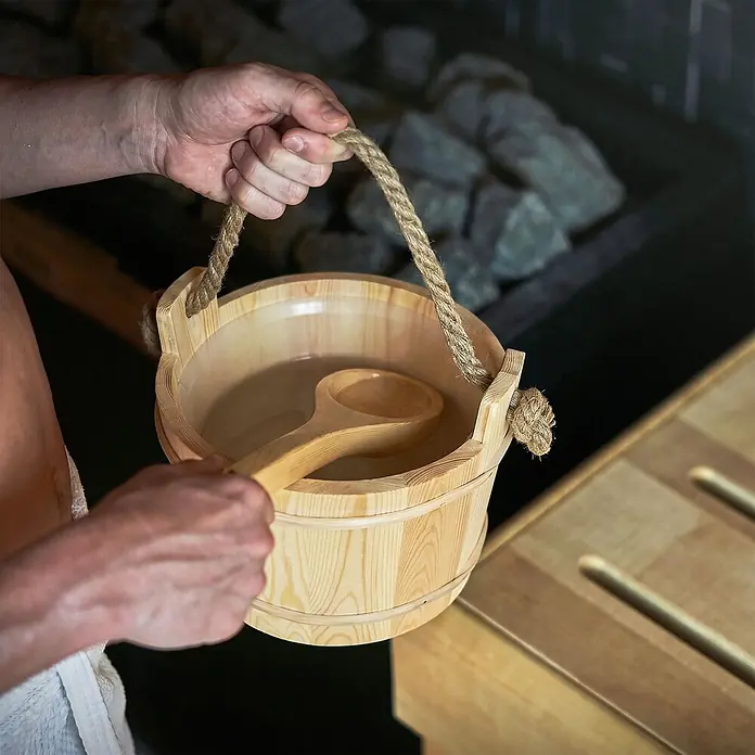 LuxeBath Saunazubehör-Set aus massivem Kiefernholz, Sauna Set 4-teilig -  - Hände halten einen hölzernen Saunaeimer mit Schöpfkelle und Wasser in einer Saunaumgebung.