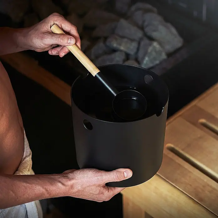 LuxeBath Sauna-Zubehör-Set - Aluminium - Person hält schwarzen Saunakübel und Schöpfkelle mit Holzgriff in der Sauna, bereit, Wasser auf heiße Steine zu gießen.