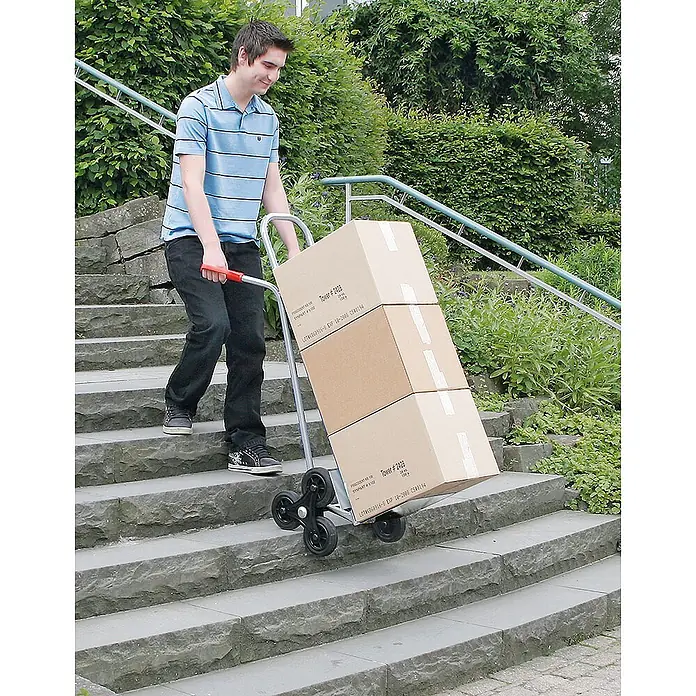BRB Treppen-Sackkarre - Aluminium - Mann transportiert Kartons eine Steintreppe hinunter mit einer metallenen Treppenkarre mit roten Griffen und schwarzen Sternrädern.