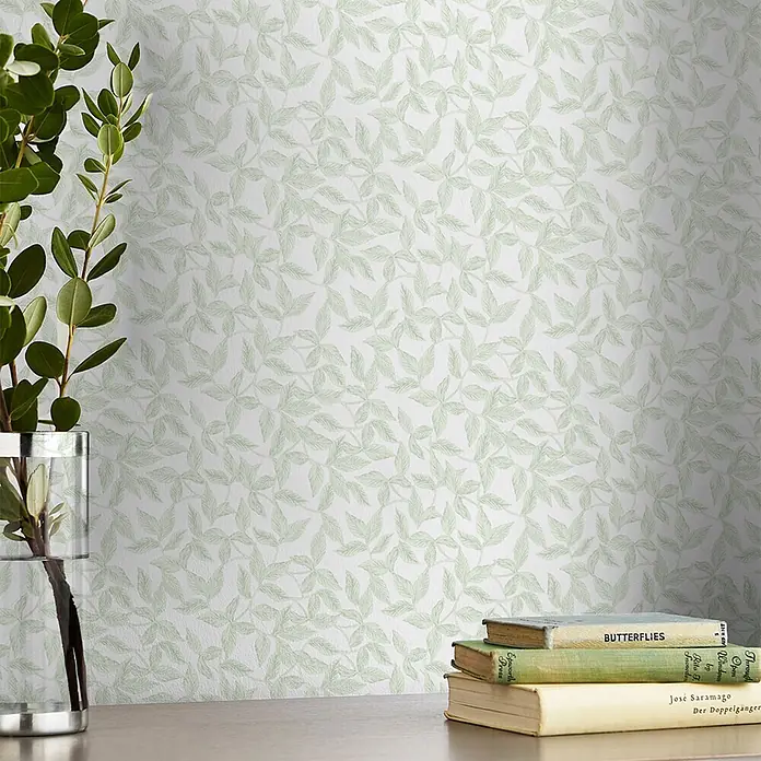 Vliestapete Erwood 1000x52cm Laura Ashley -  - Hellgrüne Tapete mit Blättermuster an einer Wand, dahinter ein Tisch mit Pflanze und Büchern.
