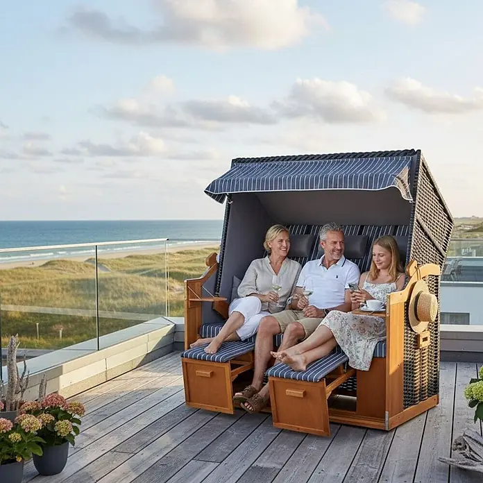 Strandkorb Ostsee XXL Wacholder PE Schwarz Dessin Royal Anthrazit -  - Familie entspannt in einem großen, geflochtenen und hölzernen Strandkorb mit blau-weiß gestreiften Kissen auf einer Terrasse mit Meerblick.