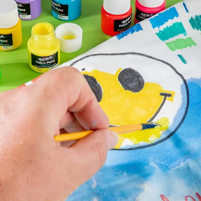 Mont Marte Textilmalfarben-Set Signature - 8x20ml - Mehrfarbig, 8 Stk. x 20 ml - Eine Hand bemalt ein gelbes Smiley-Gesicht auf Stoff mit Mont Marte gelber Stofffarbe und Pinsel. Weitere bunte Farbflaschen sind sichtbar.