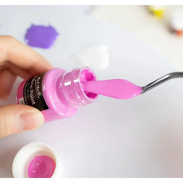 Mont Marte Textilmalfarben-Set Signature - 8x20ml - Mehrfarbig, 8 Stk. x 20 ml - Eine Hand schöpft leuchtend pinke Stofffarbe aus einer Flasche mit einem Spachtel. Die Flasche ist als 'Mont Marte Fabric Paint' beschriftet.