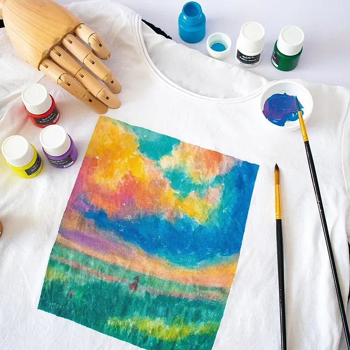 Mont Marte Textilmalfarben-Set Signature - 8x20ml - Mehrfarbig, 8 Stk. x 20 ml - Weißes T-Shirt mit einem lebendigen, handgemalten Landschaftsdesign, erstellt mit farbigen Stofffarben. Verschiedene Farbflaschen und Pinsel sind sichtbar.