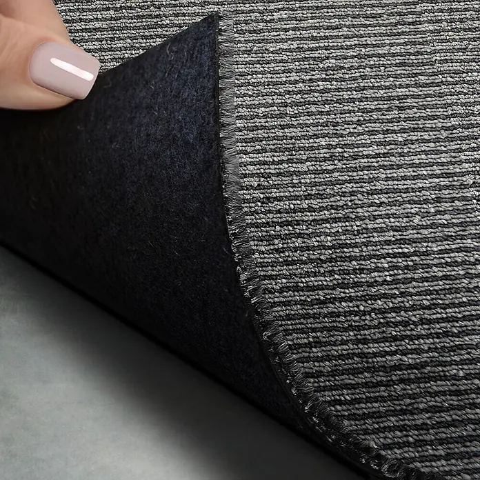 Floordirekt Teppichfliese Bohemia - 50 x 50 cm - 0,25 m² - Grau -  - Eine Hand hebt einen grau gestreiften Teppich an, der seine schwarze Filzunterseite und unfertige Kante zeigt.