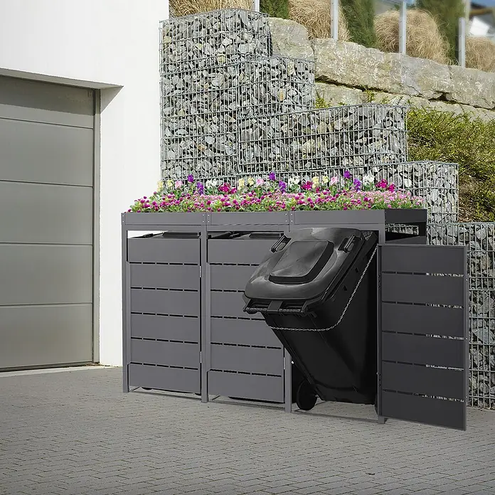Mülltonnenbox 3er mit Pflanzdach 240L 196x80x124cm Anthrazit aus Stahl -  - Dunkelgraue Lamellen-Mülltonnenbox mit Blumenbepflanzung oben, eine Tonne zum Entleeren herausgekippt, neben einer Garage platziert.