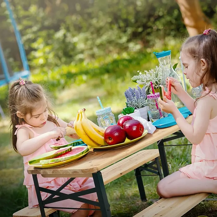 Kindersitzgarnitur Outdoor Tannenholz 2 Bänke mit Tisch Stahlgestell -  - Zwei Mädchen picknicken an einem kleinen Holztisch mit angebauten Bänken im Garten, beladen mit Obst und Getränken.