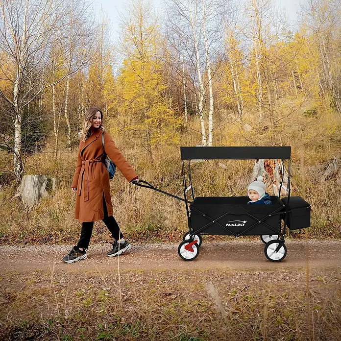 Bollerwagen faltbar mit Dach  Schwarz L für Kinder Strand Picknick -  - Schwarzer Faltwagen mit Kind, von einer Frau auf einem Herbstweg gezogen. HAUKI Logo sichtbar.