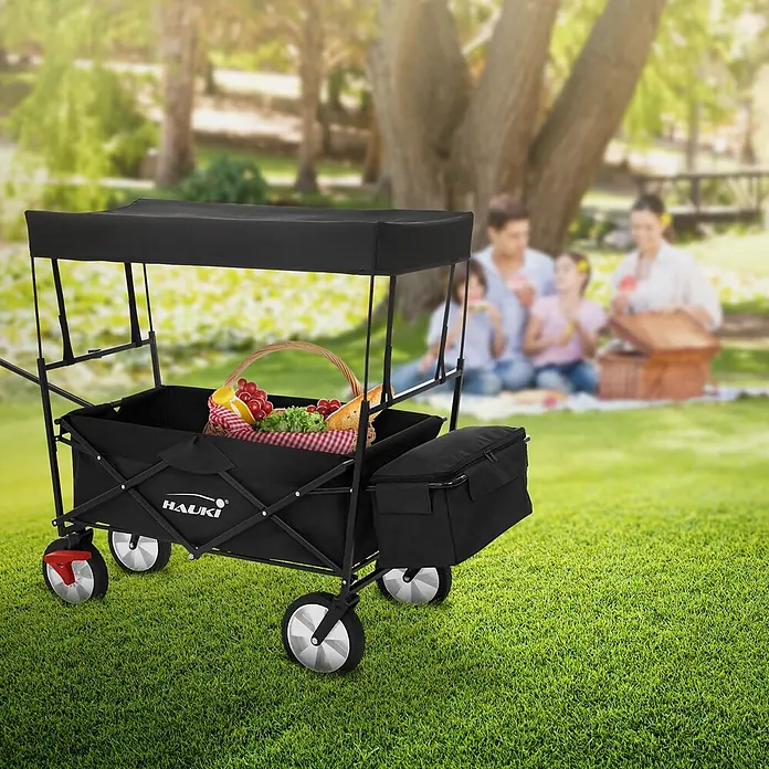 Bollerwagen faltbar mit Dach  Schwarz L für Kinder Strand Picknick -  - Schwarzer Faltwagen mit Dach und seitlicher Kühltasche, gefüllt mit Picknick-Utensilien, in einer Parkszenen mit picknickender Familie.