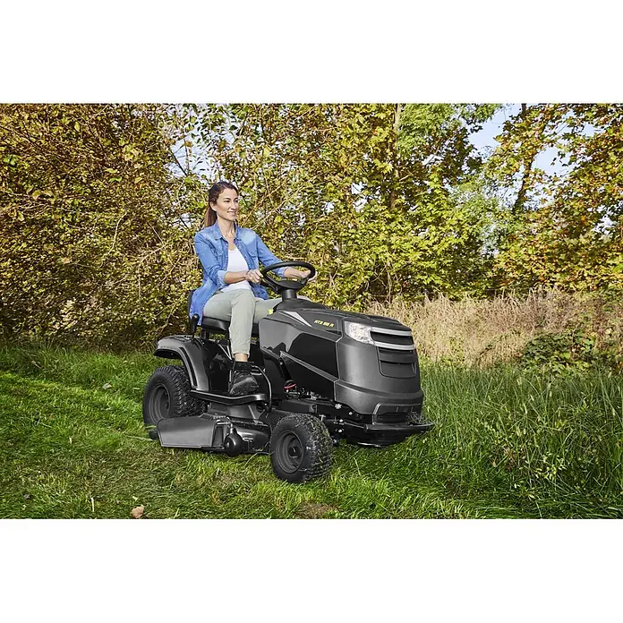 Alpina Tractor cortacésped AT3 98 A - 6,5 kW, 352 cm³, Ancho de corte: 98 cm, Recomendación máxima de superficie: 4.500 m² - Una mujer maneja un cortacésped de asiento negro y gris en una zona exterior de césped.