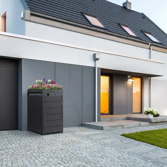 Mülltonnenbox 1er mit Pflanzdach 240L 68x80x124 cm Anthrazit aus Stahl -  - Dunkelgraue Mülltonnenbox mit Blumenbeet auf dem Dach, vor einem modernen Haus auf gepflasterter Einfahrt.