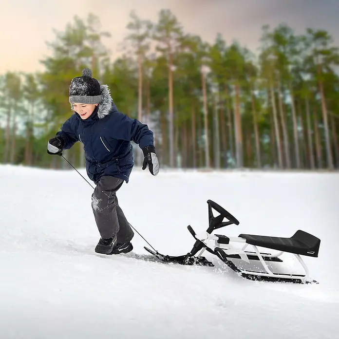 Lenkschlitten für Kinder Weiß 119 x 38 x 50 cm Stahlrahmen bis 90 kg -  - Lächelndes Kind zieht einen weiß-schwarzen Snow Racer bergauf in verschneiter Waldlandschaft. Schnee spritzt von den Kufen.