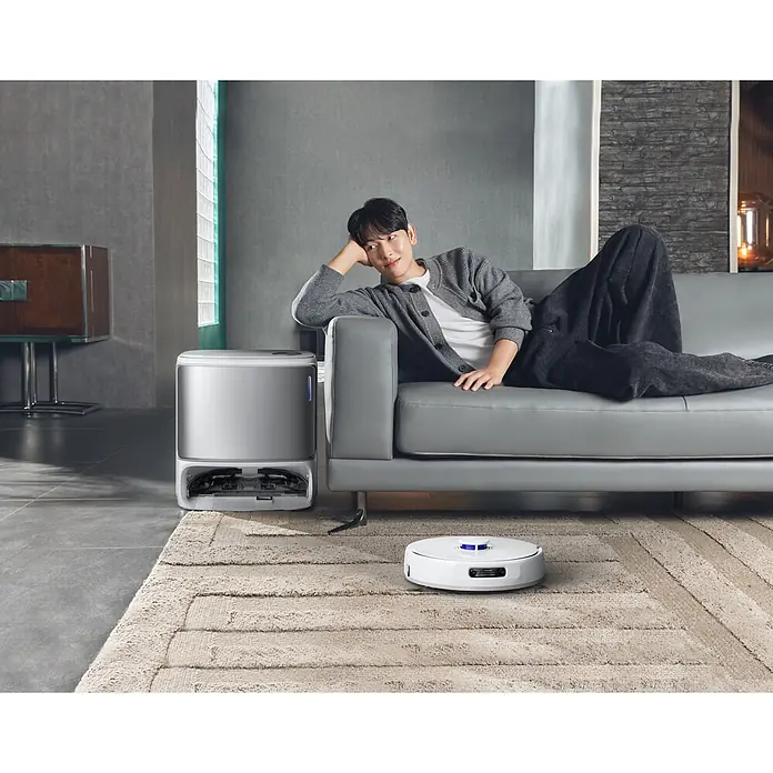 Narwal Saugwischroboter Freo Z10 ultra Extend - Saugkraft: 18.000 Pa, Akkulaufzeit: 150 min, Fassungsvermögen Roboter: 2.500 ml - Ein weißer Saugroboter reinigt einen beigen Teppich, während seine silberne Reinigungsstation neben einem Sofa steht, auf dem ein Mann entspannt.