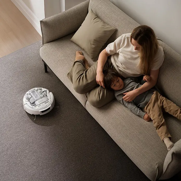 DJI Saugwischroboter A - Saugkraft: 25.000 Pa, Akkulaufzeit: 180 min, Fassungsvermögen Roboter: 260 ml - Weißer Saugroboter mit transparenter Abdeckung reinigt dunklen Teppich, während Mutter und Kind auf Sofa entspannen.