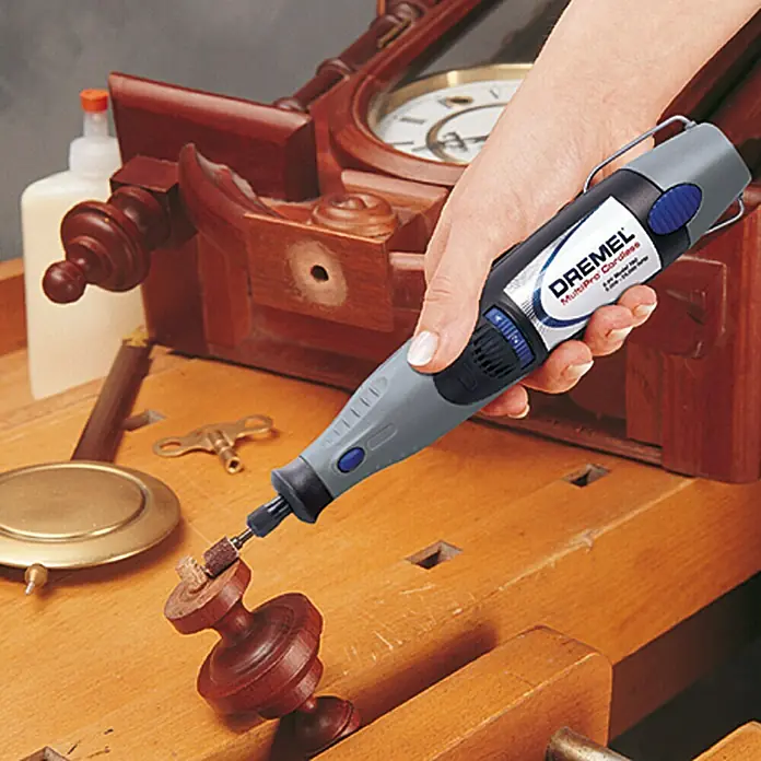 Dremel Schleifband Mod. 430 - 6,4 mm, 2 Stk., Körnung: 60 - Nahaufnahme eines Dremel MultiPro Cordless, der zum Schleifen eines Holzobjekts verwendet wird, mit einer Werkbank und einer Uhr im Hintergrund.