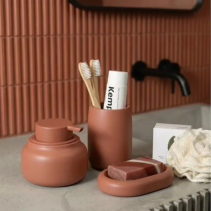 Spirella Jabonera - 8,3 x 12,7 x 2,2 mm, Terracota - Un juego de accesorios de baño de terracota, que incluye un dispensador de jabón, un portacepillos y una jabonera, sobre un mostrador de baño.