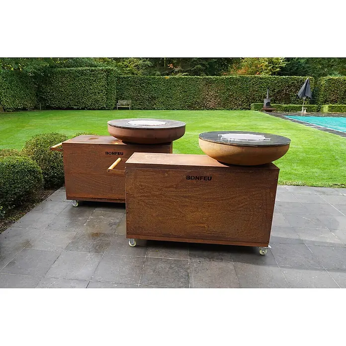 Bonfeu Brasero barbacoa Bonbiza Island - Ø x Al: 102 x 110 cm, Acero, Marrón óxido - Dos unidades de cocción exterior Bonfeu de metal oxidado con braseros, sobre bases con ruedas en un patio con jardín.