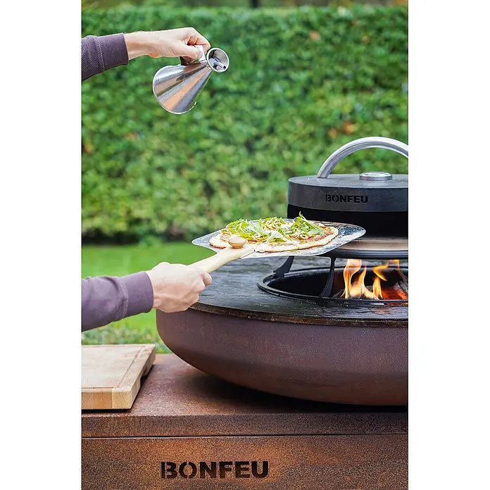 Bonfeu Brasero barbacoa Bonbiza Island - Ø x Al: 102 x 110 cm, Acero, Marrón óxido - Persona metiendo una pizza en un horno de pizza exterior negro sobre un brasero de metal rústico. Otra mano vierte aceite de un dispensador.