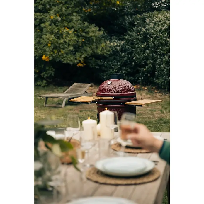 Outr Ahumador Kamado Grill Large - Diámetro de la superficie de la parrilla: 46,8 cm, Vinotinto - Parrilla cerámica roja OUTR con mesas laterales de madera en un jardín, lista para cenar al aire libre.