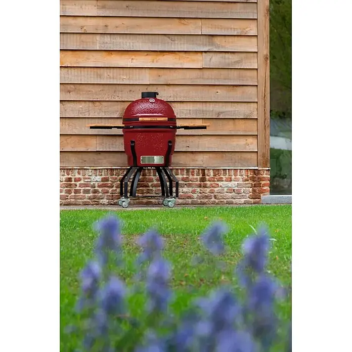 Outr Ahumador Kamado Grill Large - Diámetro de la superficie de la parrilla: 46,8 cm, Vinotinto - Parrilla de cerámica roja con estantes laterales de madera sobre un soporte con ruedas, situada al aire libre frente a una pared de madera.