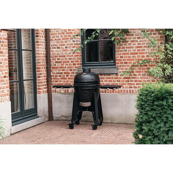 Outr Ahumador Kamado Grill Rock Large - Diámetro de la superficie de la parrilla: 46,8 cm, Negro - Parrilla kamado negra con mesas laterales en un soporte con ruedas, en un patio junto a un edificio de ladrillo.