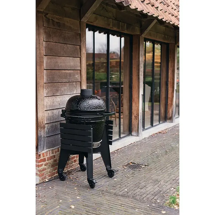 Outr Ahumador Kamado Grill Rock Large - Diámetro de la superficie de la parrilla: 46,8 cm, Negro - Parrilla kamado negra con acabado cerámico texturizado sobre un soporte rodante negro, situada al aire libre junto a un edificio rústico.