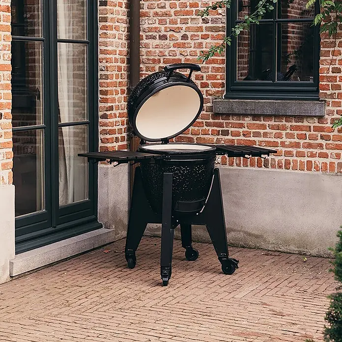 Outr Ahumador Kamado Grill Rock Large - Diámetro de la superficie de la parrilla: 46,8 cm, Negro - Parrilla kamado de cerámica negra con tapa abierta, interior crema, sobre un soporte con ruedas y estantes laterales de madera, al aire libre en un patio.