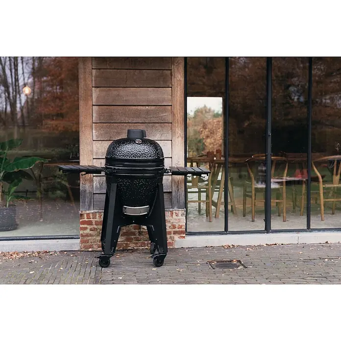 Outr Ahumador Kamado Grill Rock Large - Diámetro de la superficie de la parrilla: 46,8 cm, Negro - Parrilla kamado negra con superficie texturizada y mesas laterales sobre ruedas, situada al aire libre junto a un edificio.
