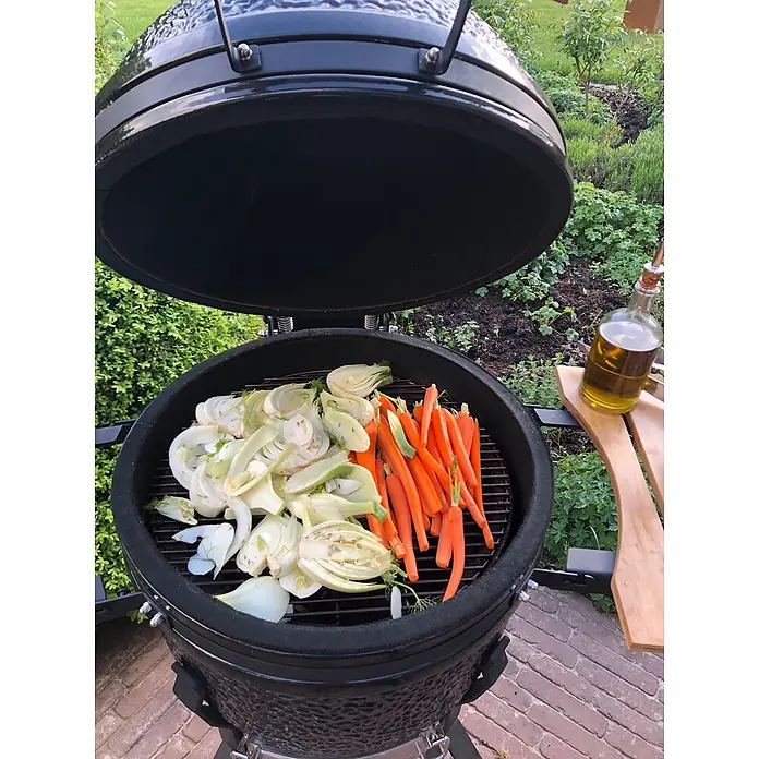 Outr Ahumador Kamado Grill Large - Diámetro de la superficie de la parrilla: 46,8 cm, Negro - Parrilla kamado de cerámica negra abierta con hinojo y zanahorias en la rejilla, lista para cocinar. Botella de aceite de oliva en el estante lateral.