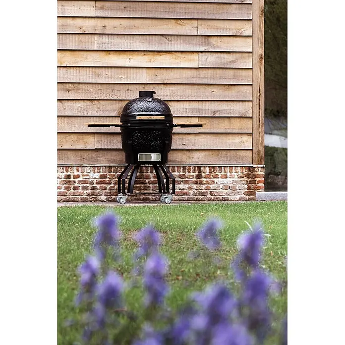 Outr Ahumador Kamado Grill Large - Diámetro de la superficie de la parrilla: 46,8 cm, Negro - Parrilla kamado de cerámica negra con mesas laterales de madera y ruedas, en exteriores frente a una pared de madera y base de ladrillo.